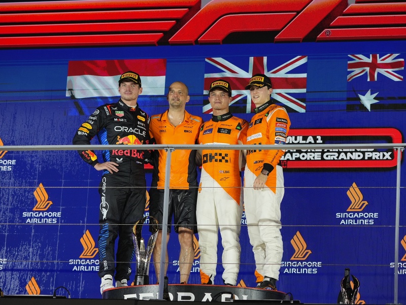 Stupeň víťazov VC Singapuru - zľava Max Verstappen, Lando Norris po boku člena tímu McLaren, a celkom vľavo Oscar Piastri