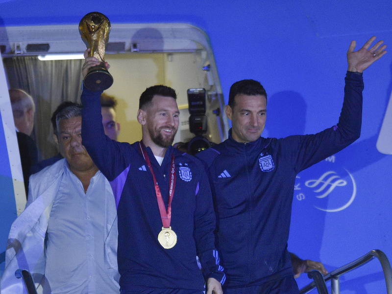 Argentínsky futbalista Lionel Messi drží trofej pre víťaza MS vo futbale počas vystupovania z lietadla, vpravo tréner Argentíny Lionel Scaloni